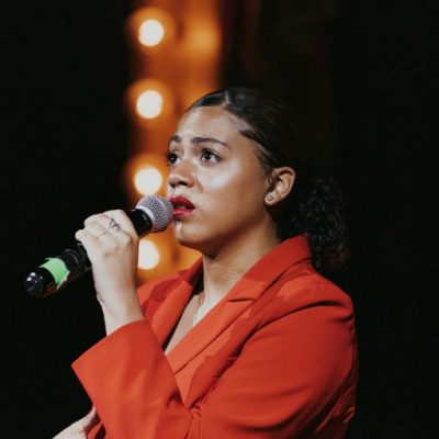 Amyah in a red jacket holding a microphone and singing.